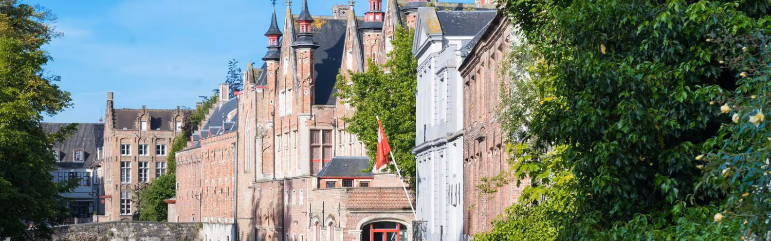 Historic red brick buildings with pointed rooftops along a canal with green trees and a clear blue sky