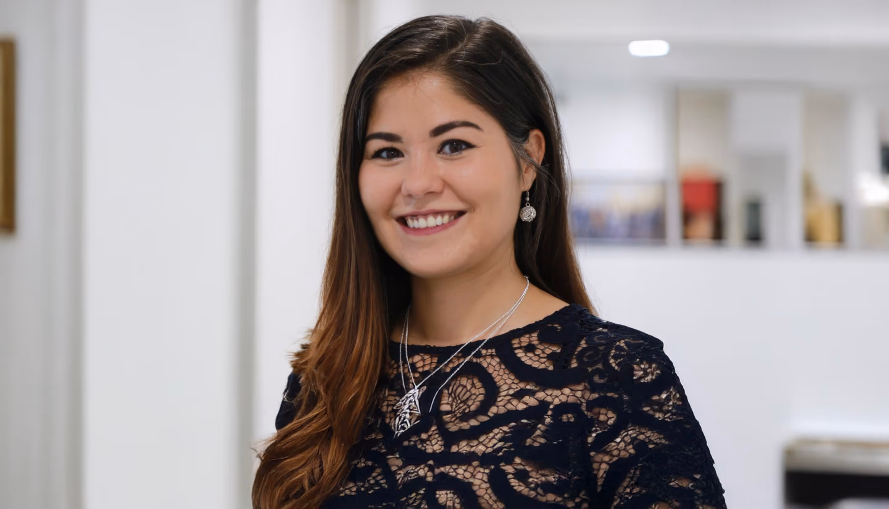 Smiling woman with long brown hair wearing a black lace top and silver jewelry in a bright indoor setting.