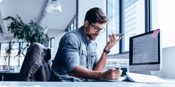 Man in glasses speaking into smartphone while taking notes at desk with computer in a bright office.